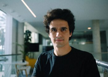 A man wearing a large black t-shirt stands in a sunlit office.