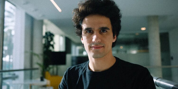A man wearing a large black t-shirt stands in a sunlit office.