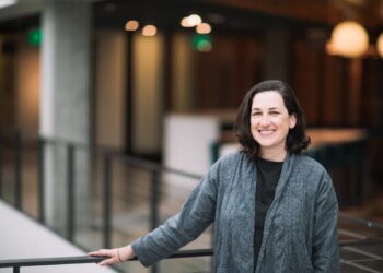 A dark-haired woman poses by a balcony in a gray cardigan sweater.