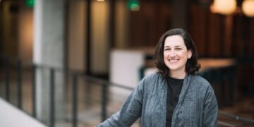 A dark-haired woman poses by a balcony in a gray cardigan sweater.