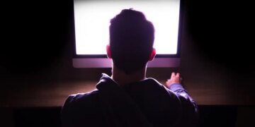 Back view of a man in front of bright computer working in the dark.